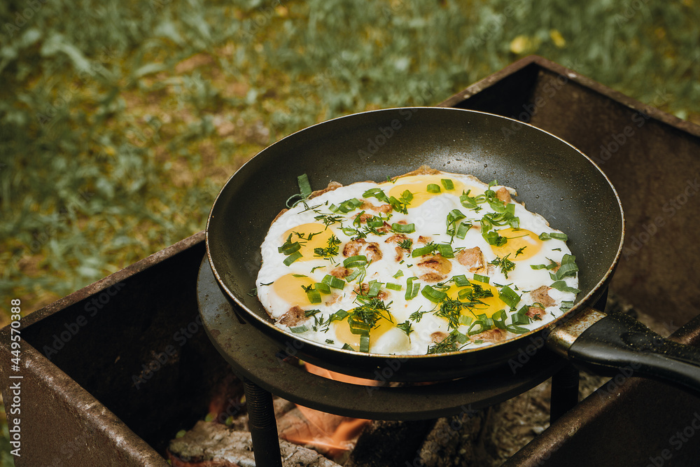Frying pan of fried eggs, bacon and organic greens, green grass on the background, cooked on the grill on a summer sunny day, summer breakfast, eating in nature - the concept of a healthy lifestyle