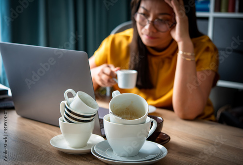A piles of used coffee cups in front of an overtired Asian woman holding a cup of coffee sitting working frustrated exhausted looking at laptop screen. Caffeine addicted bad lifestyle concept.  photo