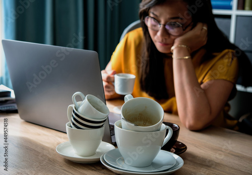 A piles of used coffee cups in front of an overtired Asian woman holding a cup of coffee sitting working frustrated exhausted looking at laptop screen. Caffeine addicted bad lifestyle concept.  photo