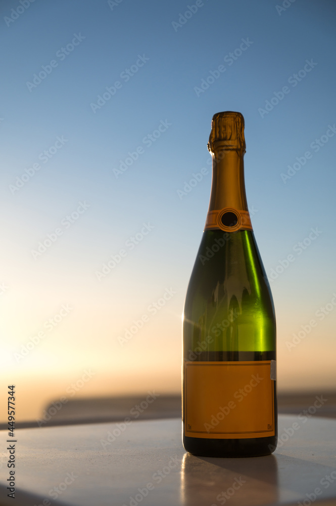 Close-up of bottle of champagne on white table on terrace. Sparkling wine. Sunset sky on background. Close-up of bottle of champagne on white table on terrace. Sparkling wine. Sunset sky on background.
