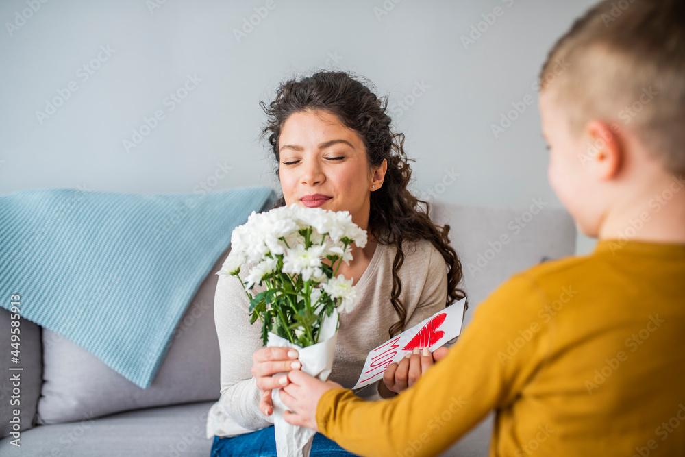 Selective focus of cute boy touching face of happy mom sitting in bed near flowers and mothers day card. Cheerful woman hugging boy and reading handmade greeting card with heart Selective focus of cute boy touching face of happy mom sitting in bed near flowers and mothers day card. Cheerful woman hugging boy and reading handmade greeting card with heart