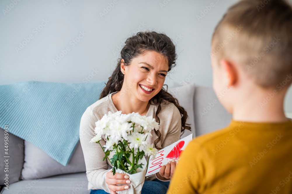 Boy hugging his mom when giving her a present and a card for mother's day. Cheerful woman hugging boy and reading handmade greeting card with heart while resting on bed Boy hugging his mom when giving her a present and a card for mother's day. Cheerful woman hugging boy and reading handmade greeting card with heart while resting on bed