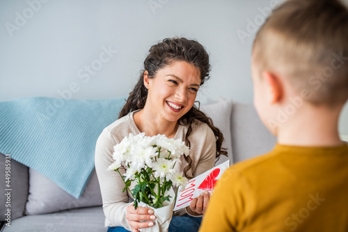 Boy hugging his mom when giving her a present and a card for mother's day. Cheerful woman hugging boy and reading handmade greeting card with heart while resting on bed