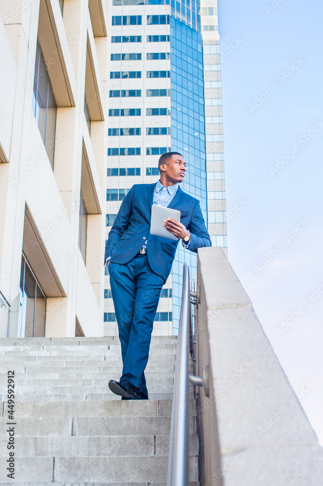 Holding a tablet computer, leaning on a railing, a young handsome black businessman is standing outside of a business building, reading, thinking and working.