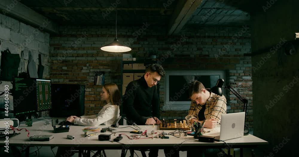 Two Men Play Chess in IT Electronics Technology Startup in Loft Office at Night while Woman Works. Corporate Environment or Male Friends Meeting For Board Game in Garage or Workshop. 4K Wide Pan Shot Two Men Play Chess in IT Electronics Technology Startup in Loft Office at Night while Woman Works. Corporate Environment or Male Friends Meeting For Board Game in Garage or Workshop. 4K Wide Pan Shot