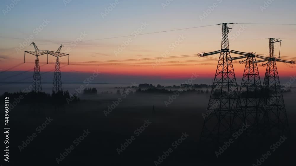 Aerial view silhouettes high voltage steel power pylons in field covered with fog countryside. Misty early morning, dawn. Drone flight low over power transmission lines. Electric tower line, sunrise