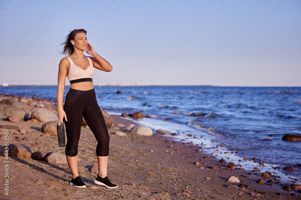 Woman in sportswear stands near seaside with bottle of water during. Woman in sportswear stands near seaside with bottle of water during.