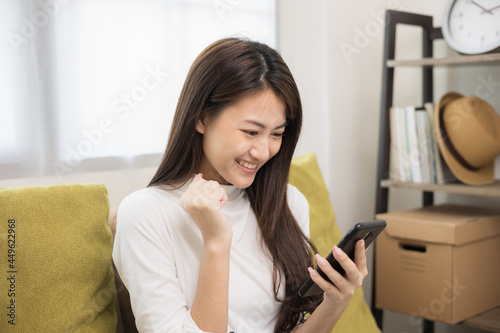 Happy young asian woman looking at screen of smartphone sitting on sofa in living room she shopping online. She very happy. Teenage girl reading news from mobile phone feeling excited.