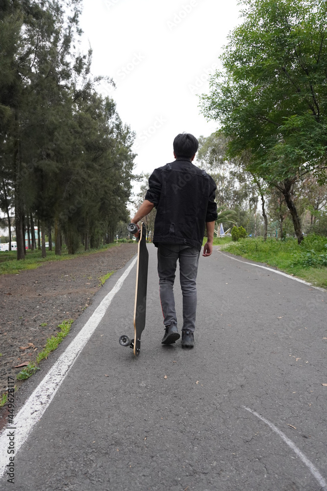 Young man with a longboard in a park. He is walking on the asphalt. Young man with a longboard in a park. He is walking on the asphalt.