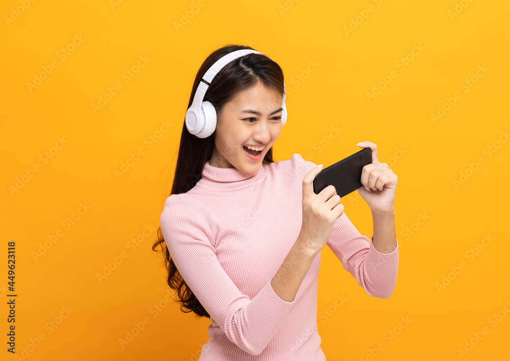 Beautiful young asian women play mobile game and put on wireless headphone standing on isolated yellow background. Playing game on smartphone winning victory moment. Very enjoy and fun relax time Beautiful young asian women play mobile game and put on wireless headphone standing on isolated yellow background. Playing game on smartphone winning victory moment. Very enjoy and fun relax time