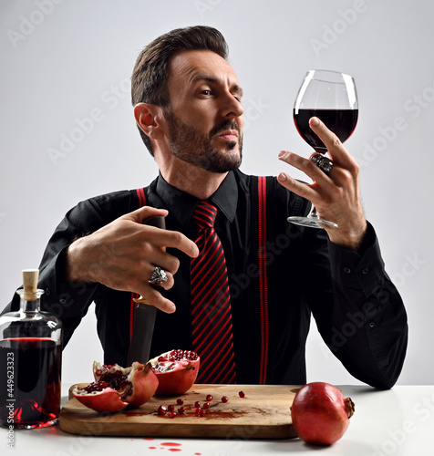 Stylish man expert, sommelier in black shirt with striped tie and suspenders sits at table with fresh pomegranate, looking at glass of freshly squeezed juice he holds in hand with one eye narrowed 