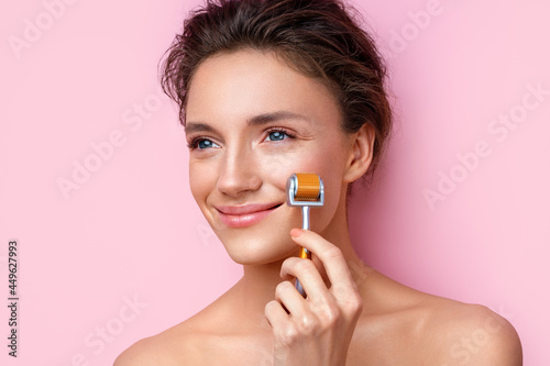Beautiful woman using derma roller for her facial skin. Photo of woman on pink background. Beauty and skin care concept photo