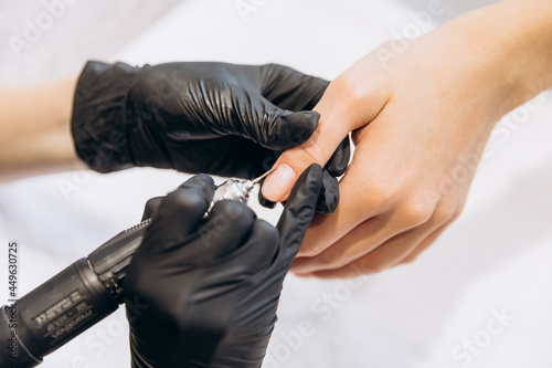 Women's hands in black gloves hold other hands and work with a manicure machine with a round-shaped nozzle on the nail. Creating a manicure in the salon as an option for rest and relaxation. © anisha_mur