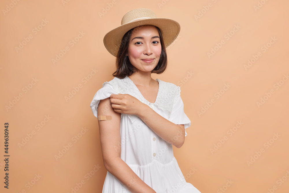 Portrait of good looking Asian woman with dark hair shows arm with adhesive plaster after getting inoculation of vaccine wears fedora and white dress isolated over beige background. Health care Portrait of good looking Asian woman with dark hair shows arm with adhesive plaster after getting inoculation of vaccine wears fedora and white dress isolated over beige background. Health care