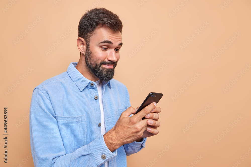 Pleased bearded man smiles happily while reading message received on smartphone wears blue velvet shirt stands against beige background copy space for your advertisement chats online with friends Pleased bearded man smiles happily while reading message received on smartphone wears blue velvet shirt stands against beige background copy space for your advertisement chats online with friends