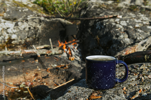 Metal cup with tea and bowler on the campfire in background. Morning in a camp. Camping and hiking concept.