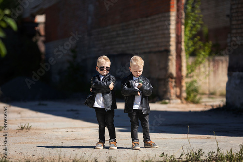 Blond twin boys in dark glasses and leather jackets.The little twins are stylishly dressed in a rock style.Cute kids look like celebrities.A children's photo on the background of an abandoned building