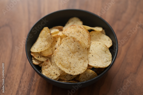 Organic potato chips with black pepper in black ceramic bowl on walnut wood background
