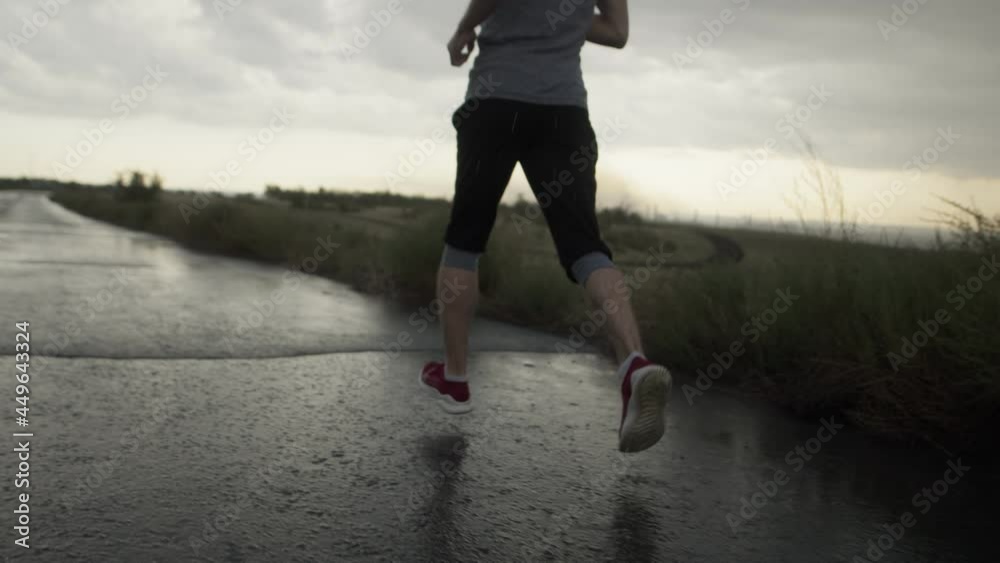 Serious man of athletic build runs through rain on road against background of cloudy gray sky and fields, back and down view. Sports motivation
