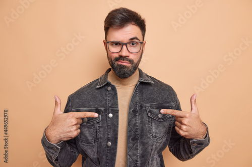 Confident serious bearded adult man makes choose me gesture indicates with index fingers at himeself feels self assured wears glasses black denim jacket poses indoor. Self promoting concept. photo