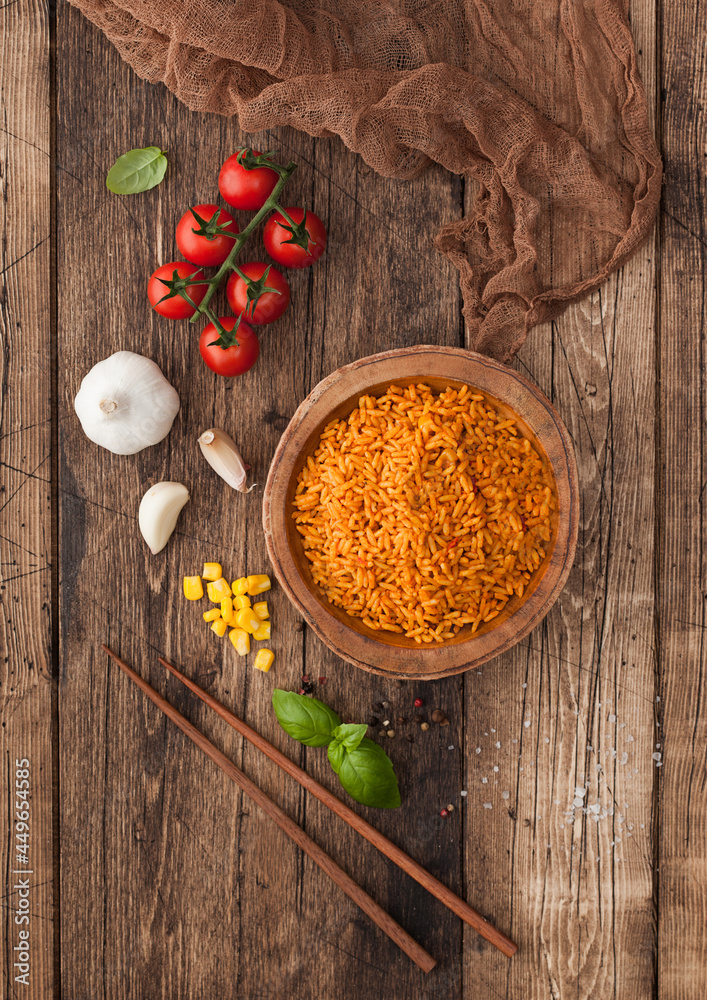 Wooden bowl with boiled red long grain basmati rice with vegetables on wooden table background with sticks and tomatoes with corn,garlic and basil. Top view