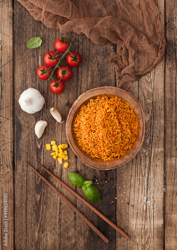Fototapeta Naklejka Na Ścianę i Meble -  Wooden bowl with boiled red long grain basmati rice with vegetables on wooden table background with sticks and tomatoes with corn,garlic and basil. Top view