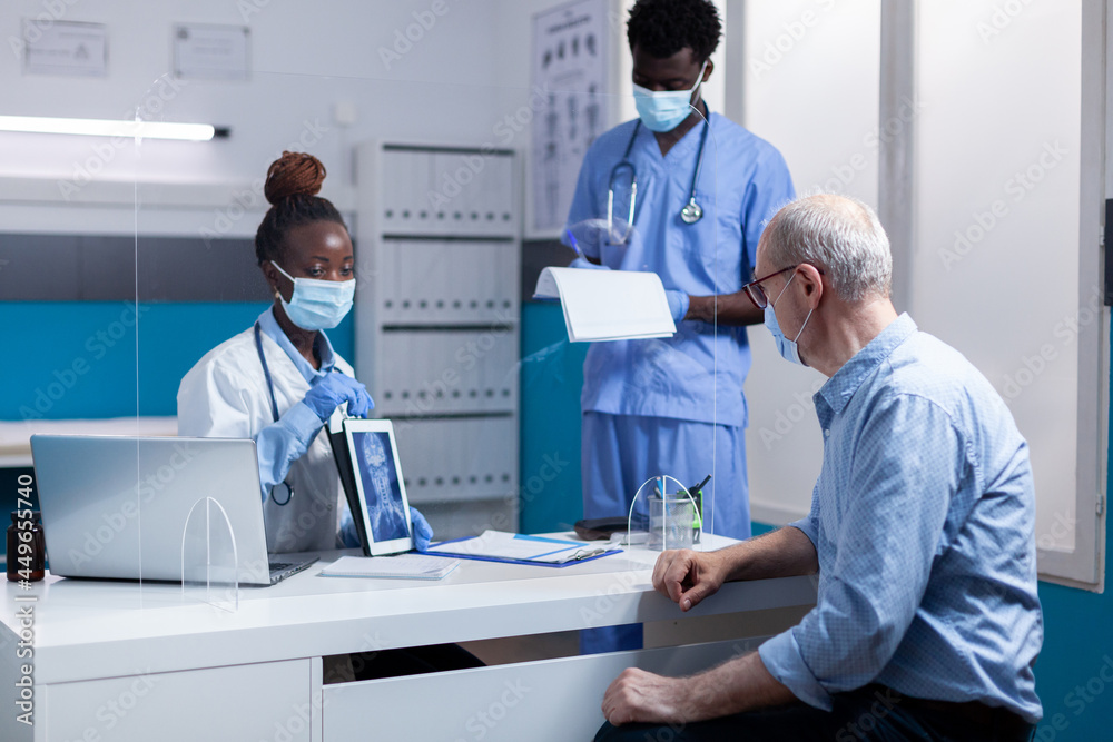 Obraz premium Medical team of black people talking to elderly patient sitting at desk office. African american doctor holding x ray scan on digital tablet showing diagnosis to senior man with disease