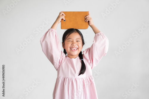 Asian child or kid girl smile laughing holding and show computer tablet or smartphone pad on overhead to learn from home or study online and education communication on white gray background isolated