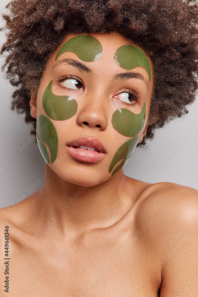Vertical shot of thoughtful African American woman looks away thoughtfully applies green hydrogel patches on face for skin care stands shirtless has mysterious expression. Beauty treatments. Vertical shot of thoughtful African American woman looks away thoughtfully applies green hydrogel patches on face for skin care stands shirtless has mysterious expression. Beauty treatments.