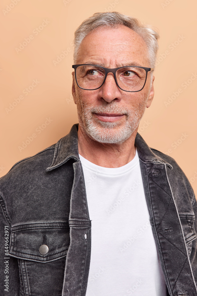 Vertical shot of serious grey haired man with beard concentrated away wears transparent glasses black denim jacket thinks deeply about something isolated over beige background. People and thoughts