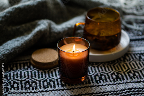 Cozy burning candle in brown glass jar, winter home interior decor with cup and blanket photo