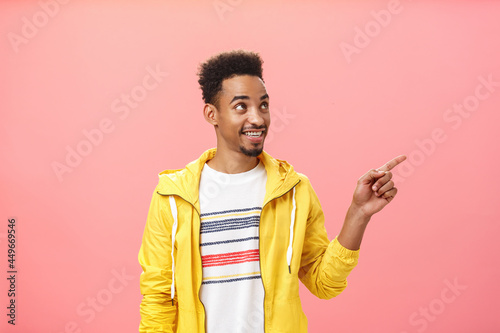 Indoor shot of intrigued and pleased african american male student in trendy yellow jacket pointing and gazing at upper right corner with dreamy delighted look enjoying observing cool display