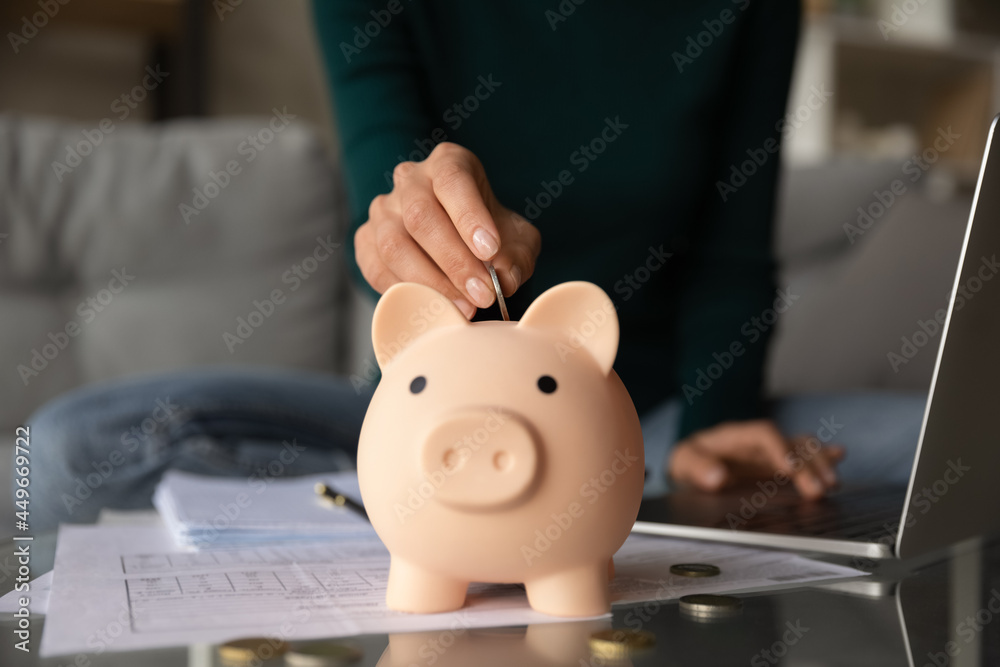 Close up woman putting coin into piggy bank, investment or insurance concept, young female using laptop, calculating domestic bills, checking financial documents, receipts, planning budget Close up woman putting coin into piggy bank, investment or insurance concept, young female using laptop, calculating domestic bills, checking financial documents, receipts, planning budget