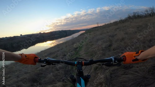The cyclist rides along the trail along the river towards the sunset. First-person view.