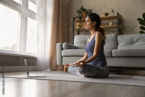 Mindful woman meditating, practicing yoga online at home, using laptop, involved in internet lesson, class with trainer, calm young female with closed eyes relaxing, breathing deep, sitting on floor