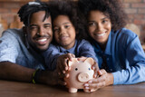 Close up blurred portrait of smiling young African American family with daughter recommend saving money in piggybank. Happy ethnic parents with small child feel economical make financial investment.