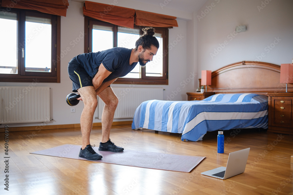 Bearded man doing kettlebell swing to workout at home with online class on laptop. Bearded man doing kettlebell swing to workout at home with online class on laptop.