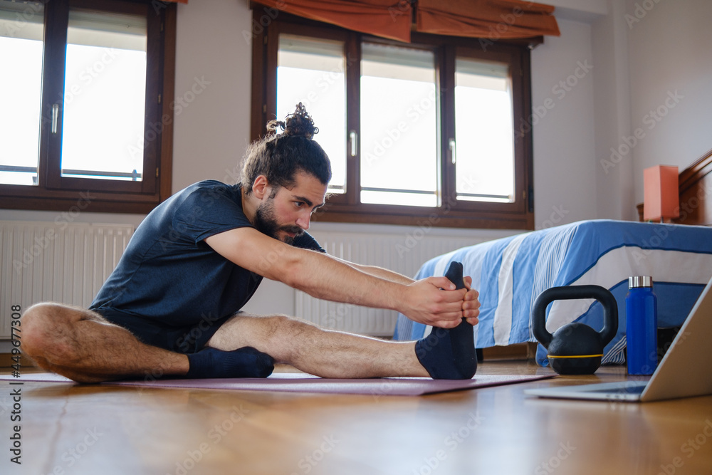 Fototapeta premium Man doing deep stretches at home watching an online exercise class on laptop.