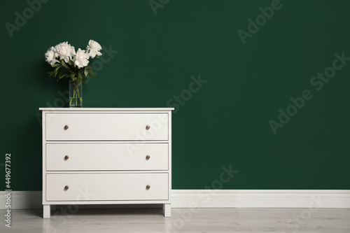 Fototapeta Naklejka Na Ścianę i Meble -  Modern white chest of drawers with beautiful bouquet near green wall indoors. Space for text