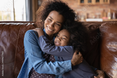 Happy loving young African American mother and small teen daughter relax on sofa hug cuddle at home together. Smiling caring ethnic mom and little biracial girl child embrace. Motherhood concept.