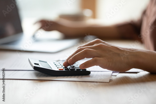 Close up of Indian woman manage household budget finances count on calculator make payment on computer. Ethnic female calculate expenses expenditures on machine pay bills on laptop online.