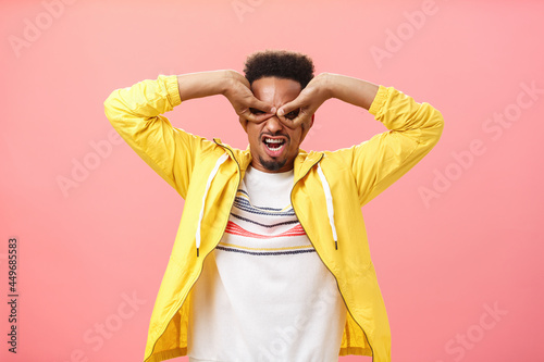 Man from childhood dreamt become superhero. Childish playful african american young guy acting immature making mask from hands over eyes aping standing in yellow jacket over pink wall photo