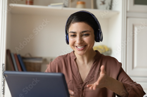Smiling young Indian woman wear earphones use tablet have webcam digital virtual event on gadget. Happy teen female in headphones speak talk on video call on pad device. Communication concept.