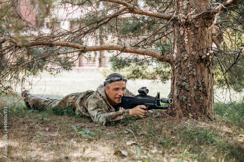 Man is lying on the green grass and looking into the optical sight a weapon. People playing laser tag shooting game in outdoor. War simulation game