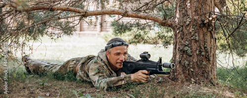 Man is lying on the green grass and looking into the optical sight a weapon. People playing laser tag shooting game in outdoor. War simulation game. Banner photo