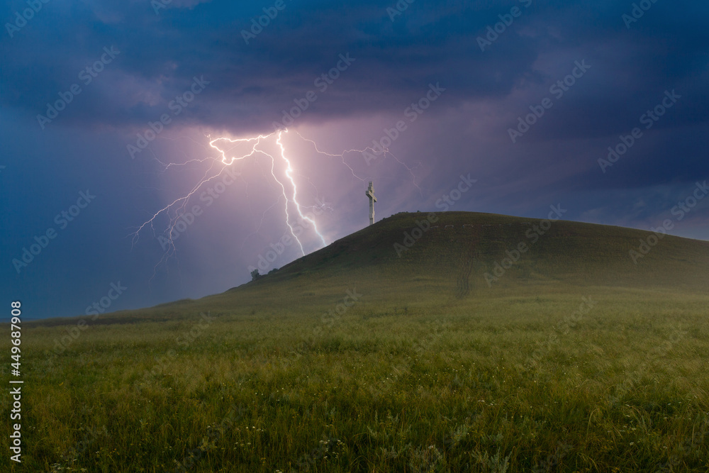 Summer awe landscape with flashes of bright lightning on dark cloudy sky during a thunderstorm over the Orthodox cross at the top of the hill. Drokinskaya mountain near Krasnoyarsk, Russia Summer awe landscape with flashes of bright lightning on dark cloudy sky during a thunderstorm over the Orthodox cross at the top of the hill. Drokinskaya mountain near Krasnoyarsk, Russia