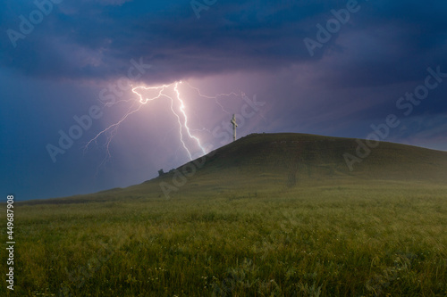 Summer awe landscape with flashes of bright lightning on dark cloudy sky during a thunderstorm over the Orthodox cross at the top of the hill. Drokinskaya mountain near Krasnoyarsk, Russia