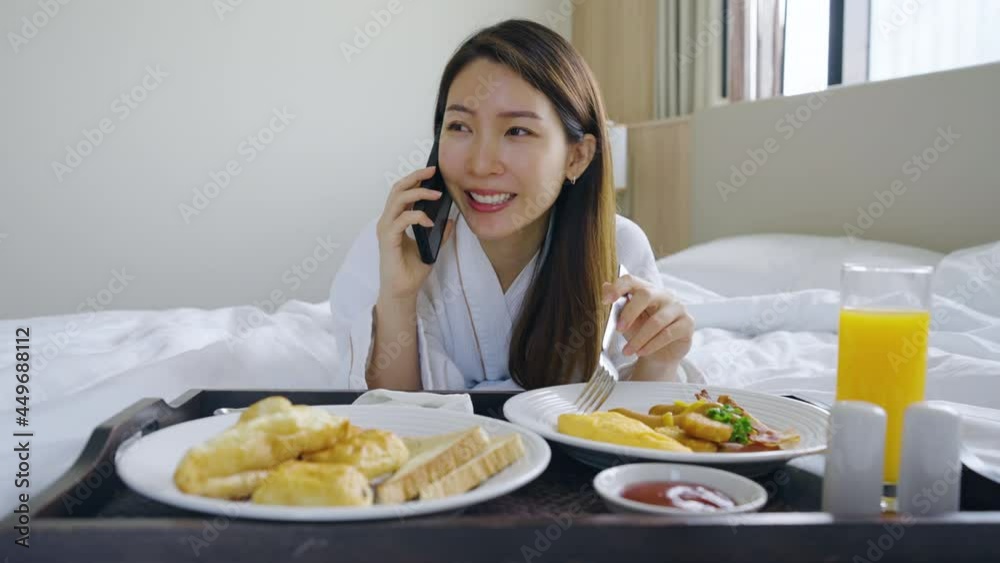Portrait beautiful young Asian woman wearing bathrobe talking on a phone while eating breakfast on a bed in bedroom Portrait beautiful young Asian woman wearing bathrobe talking on a phone while eating breakfast on a bed in bedroom
