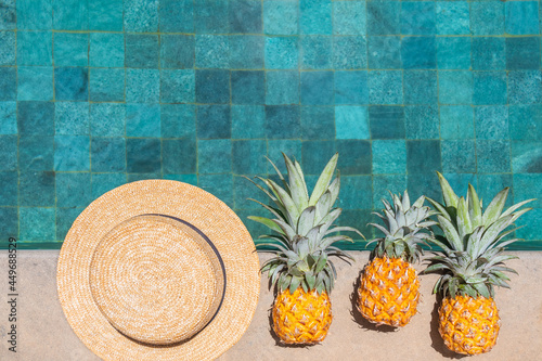 A hat and three pineapples are in a row by the water. Top view, copy space.