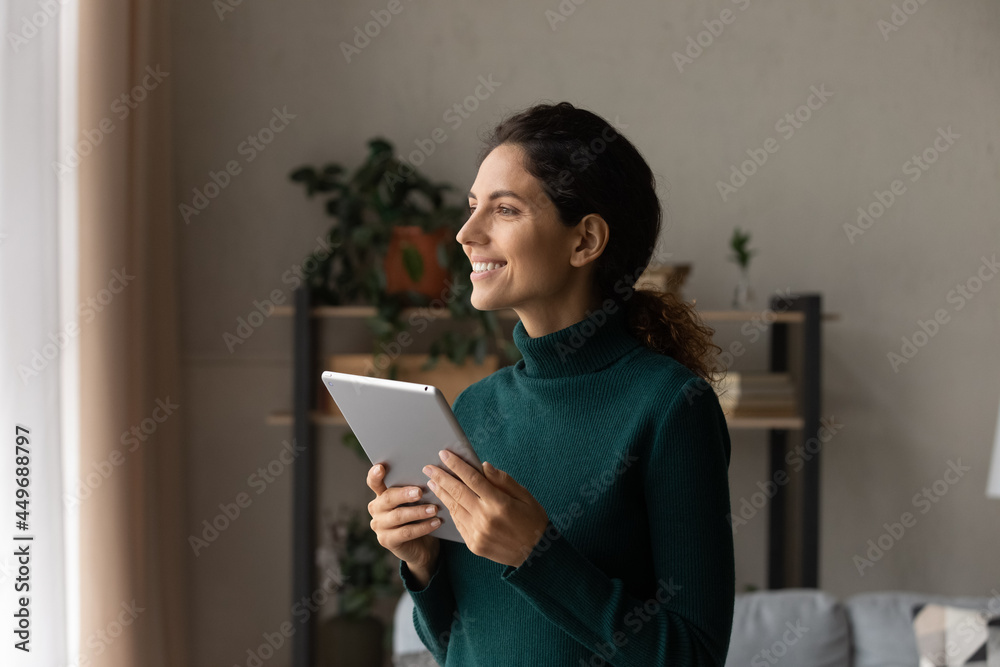 Smiling dreamy woman distracted from tablet, looking out window, standing at home, visualizing good future, excited by great news, attractive female chatting online with boyfriend or waiting for call Smiling dreamy woman distracted from tablet, looking out window, standing at home, visualizing good future, excited by great news, attractive female chatting online with boyfriend or waiting for call